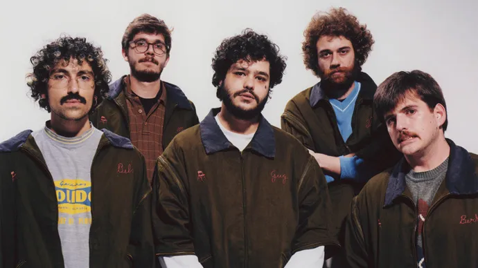 Cinco homens brancos, integrantes da banda Jovem Dionisio, em frente a um fundo branco.