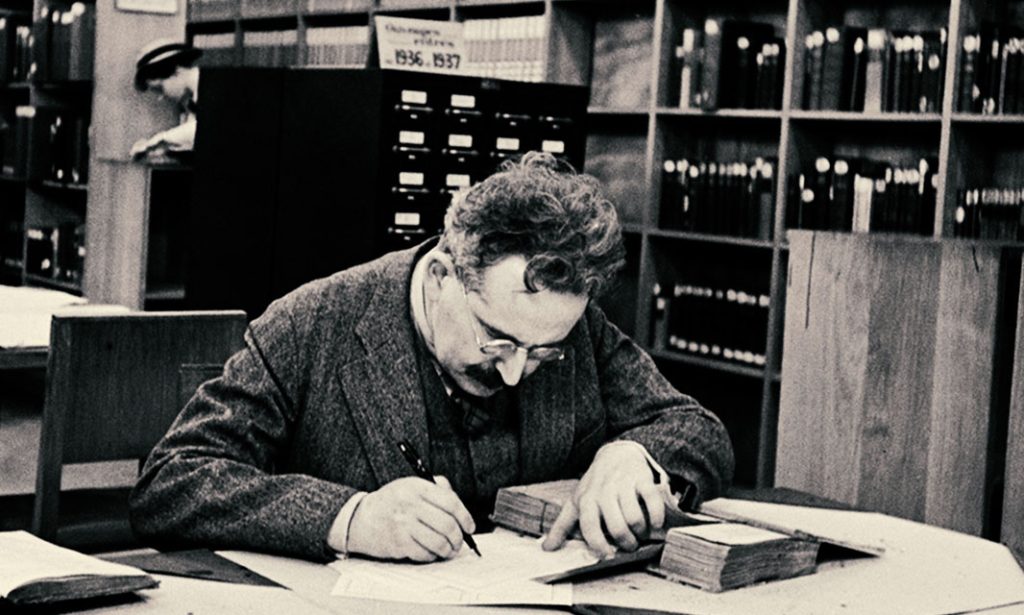 Walter Benjamin sentado em uma biblioteca, escrevendo em papéis sobre a mesa.